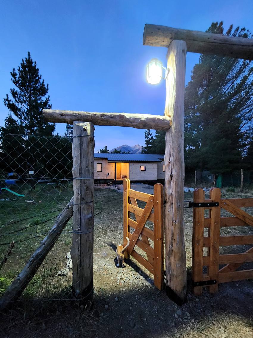La tranquera de entrada al anochecer, con un gato y la cabaña iluminada al fondo